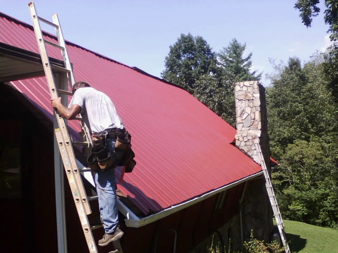 Roofer on a ladder beside a red metal roof and stone chimney — Soffit & Fascia Repair by Roofing Brothers in Broadway